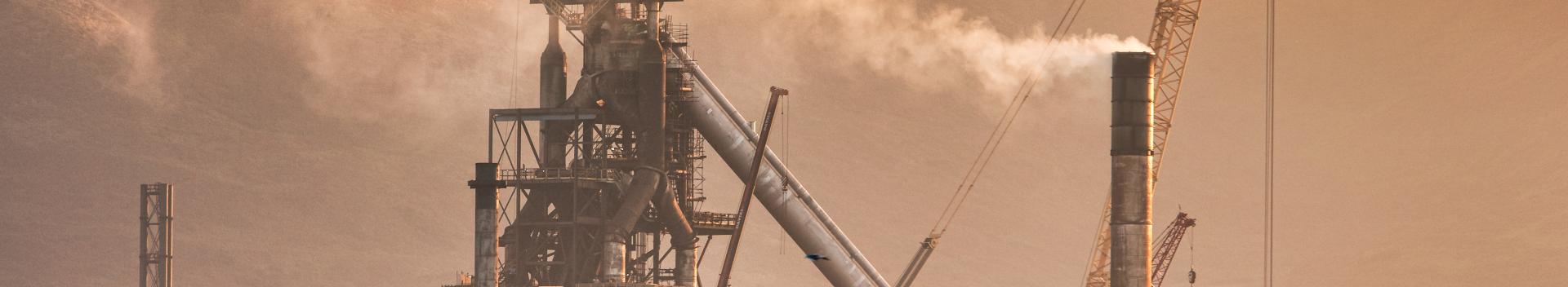 Blast furnace in the forefront with wind turbines sitting on top of the mountain in the background