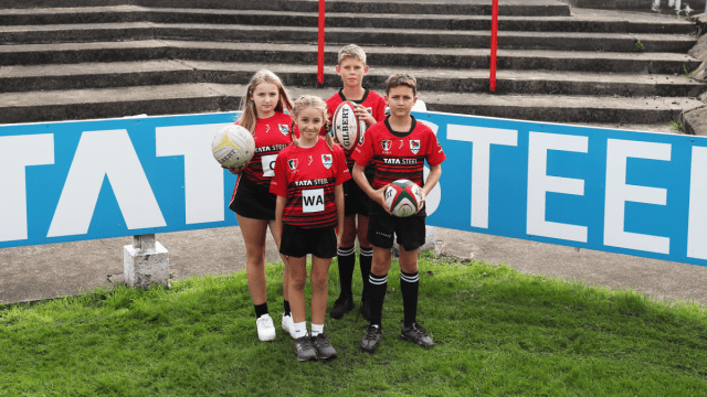 Two young girls (left) and two young boys (right) stand facing the camera on a grassy pitch dressed in red and black Tata Steel branded sports kit. The tallest girl (behind) holds a netball and the two boys on the right hold rugby balls. Light marine Tata Steel branding can be seen in the background.